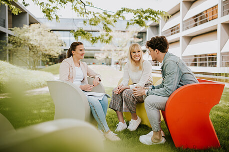 Drei junge Menschen sitzen im Campusgarten und unterhalten sich.
