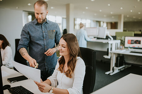 Ein Mann und eine am Schreibtisch sitzende junge Frau schauen gemeinsam auf einen Ausdruck. 