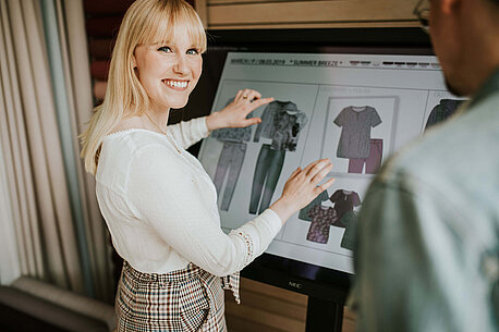 Eine blonde Frau steht vor einem Bildschirm mit Modeskizzen und lächelt in die Kamera.
