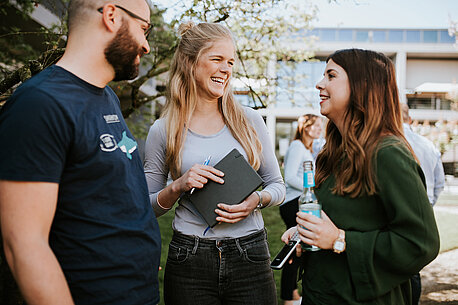 Drei Kolleg*innen stehen gemeinsam in der Gartenanlage auf dem Ernstings's family-Campus und unterhalten sich lachend.