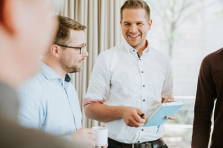 Ein Mann mit Notizbuch und Stift und ein Mann mit einem Kaffee in der Hand unterhalten sich.